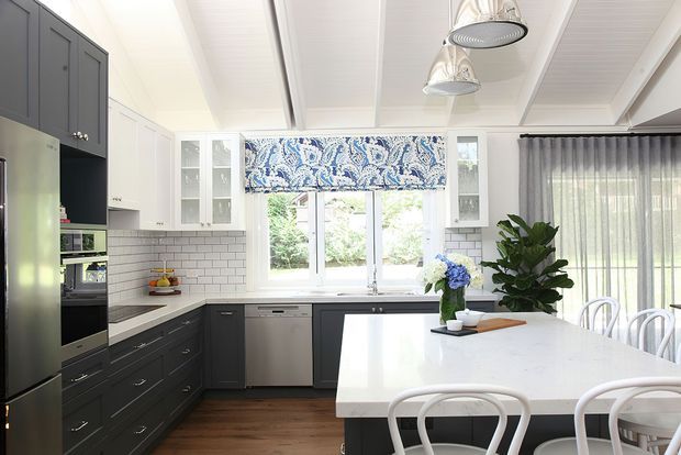 Bright kitchen and dining area with dark charcoal cabinetry, white subway-tile splashback, white stone island bench, stainless-steel appliances, and a large central window dressed with a blue patterned Roman blind. The room has a white vaulted panelled ceiling with pendant lights, timber flooring, and a glazed dining area with sheer curtains.