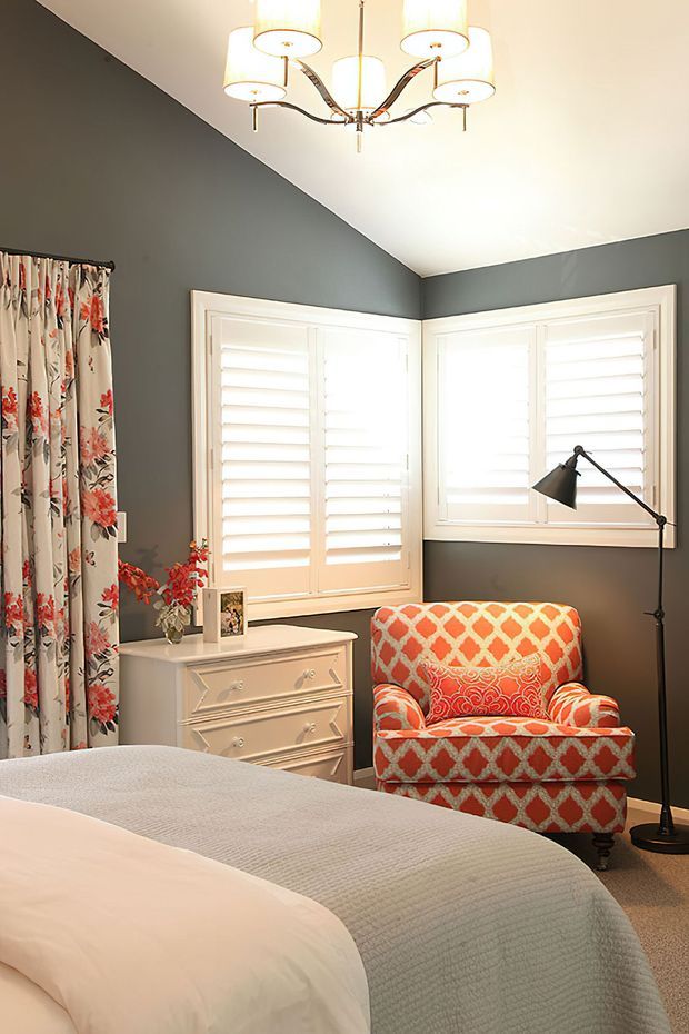 A warmly lit bedroom with dark grey walls, white plantation shutters, and a floral curtain. The room features a white decorative dresser, an orange-and-cream patterned upholstered armchair with a coordinating cushion, a black floor lamp, a chandelier, and a neatly made bed with a pale textured coverlet.
