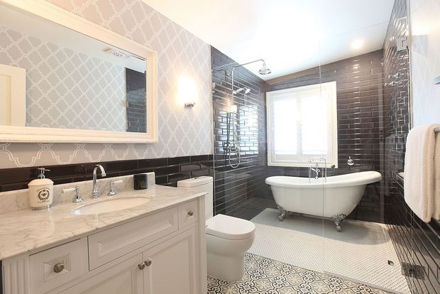A bright, traditionally styled bathroom with a freestanding clawfoot bathtub, white toilet, and a pale built-in vanity topped with marble. Glossy dark subway tiles cover the lower walls and shower area, while patterned grey wallpaper and a decorative patterned floor tile add visual detail. A framed mirror, chrome fixtures, wall sconces, and a heated towel rail complete the space.