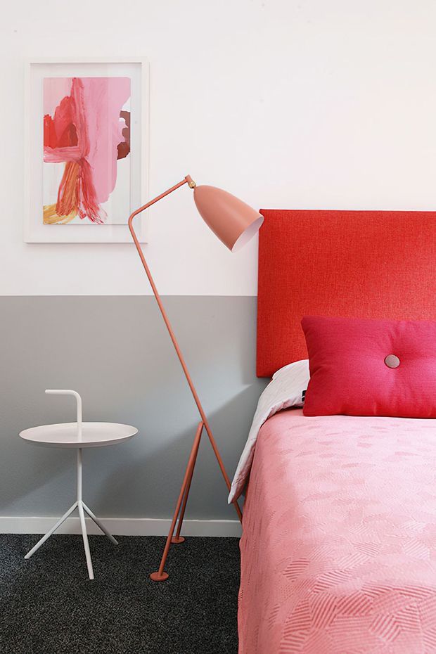 A brightly styled bedroom corner with a red upholstered headboard, pink bedding and accent cushion, a coral floor lamp, white side table, grey lower wall, white upper wall, framed abstract artwork, and dark carpet flooring.