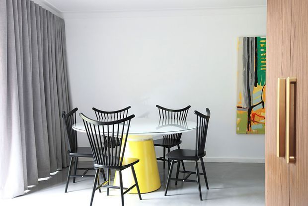 A bright, contemporary dining room with a round white dining table surrounded by black spindle-back chairs and a distinctive yellow pedestal base. Full-height grey curtains cover the left wall, while a colorful abstract artwork and timber-toned built-in joinery or door frame the right side. The room has white walls, a light grey floor, and a clean, graphic interior aesthetic.