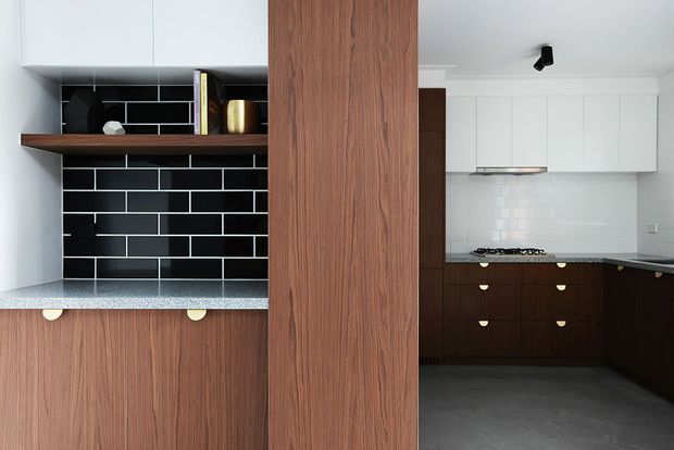 A contemporary kitchen featuring dark timber cabinetry and a central timber-clad structural divider. On the left, open shelving is set against a glossy black rectangular-tile backsplash above a speckled benchtop. On the right, the main cooking area has dark lower cabinetry, white upper cabinets, a white subway-tile backsplash, a gas cooktop, and a grey floor.