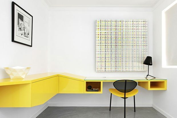 Bright minimalist home office with a vivid yellow built-in corner desk and cabinetry, paired with a black-and-yellow chair. White walls with simple ceiling moulding, grey tiled flooring, framed black-and-white artwork, a graphic grid artwork, and a modern black desk lamp create a clean mid-century-influenced workspace.