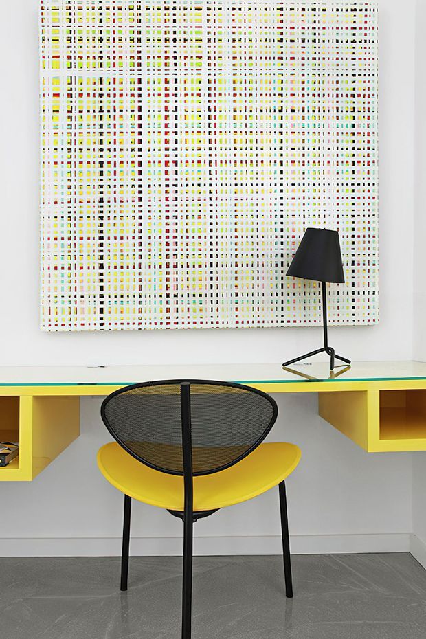A brightly styled compact workspace or console area with a vivid yellow built-in desk, a black wire-frame chair with yellow seat, and a minimalist black table lamp. A large colorful geometric grid artwork dominates the white wall, while the floor has a dark grey finish. The overall composition is graphic, playful, and mid-century inspired.