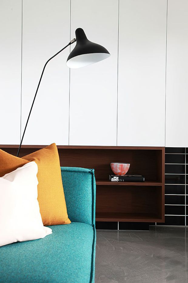 A close-up of a brightly styled modern living room featuring a vivid turquoise upholstered lounge chair or sofa with mustard-yellow and white cushions. A slim black floor lamp stands against white vertical wall panelling, while dark timber built-in shelving and a black tiled wall create contrast. The floor has a dark grey tile or stone finish, with a small book and decorative bowl displayed on the shelving.