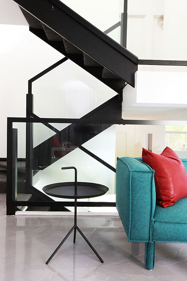 A bright contemporary interior focused on a sculptural black staircase with dark carpeted treads, geometric black balustrade framing, and clear glazing. A turquoise upholstered sofa with vivid red cushions occupies the foreground beside a slim black tray side table. The polished pale floor, white walls, and abundant daylight create a crisp, high-contrast setting.