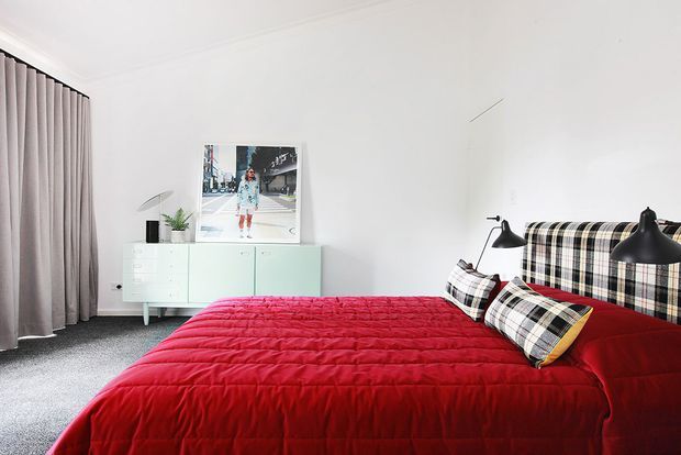 Bright contemporary bedroom with a large bed dressed in a vivid red quilt, a distinctive black-and-white plaid upholstered headboard and matching accent pillows. The room has white walls and ceiling, grey carpet, full-height taupe curtains, black bedside lamps, and a pale mint built-in sideboard beneath framed artwork.
