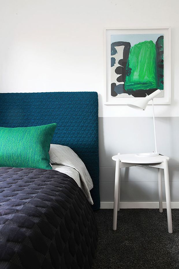 A contemporary bedroom corner with a dark blue quilted upholstered bedhead, textured dark bedding, bright teal-green cushion, white side table, and minimalist white desk lamp. White walls feature grey lower wall panelling and a framed abstract artwork with bold green and charcoal forms. Dark carpet grounds the room.