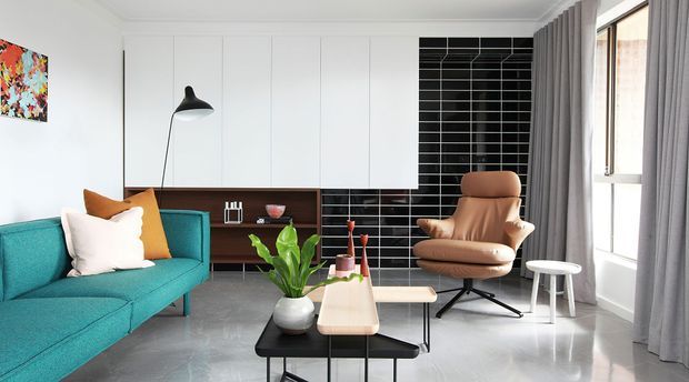 A bright contemporary living room with a turquoise sectional sofa, sculptural brown lounge chair, pale wood and black coffee tables, white built-in cabinetry, a dark timber media console, and a striking black rectangular tiled feature wall. Full-height windows with grey curtains bring in abundant natural light, while polished grey flooring and restrained decor create a refined mid-century-inspired interior.