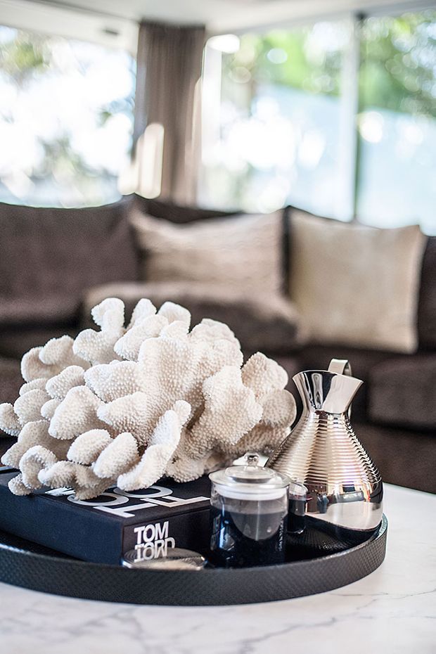 Close-up interior vignette of a living room with a dark upholstered sofa, neutral cushions, and large windows overlooking greenery. A black tray on a light marble coffee table holds a white coral-like decorative object, a reflective silver carafe, a glass jar, and a design book. The image has a bright, softly blurred, contemporary styling.