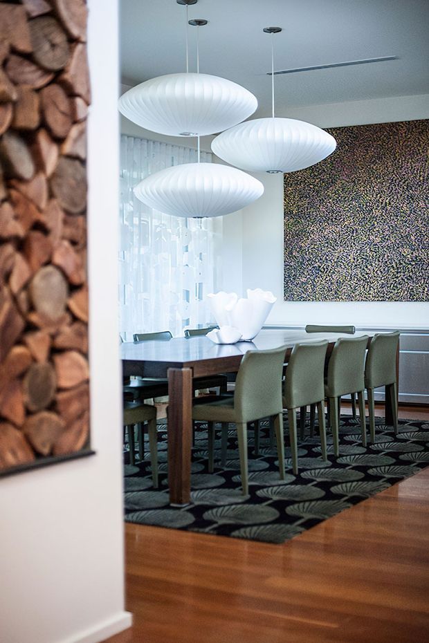 A refined dining room viewed from an adjoining space, featuring a long dark dining table with upholstered chairs, three large sculptural pendant lights, timber flooring, sheer curtains, and a large abstract wall artwork. A textured wood-slice artwork is partially visible in the foreground.