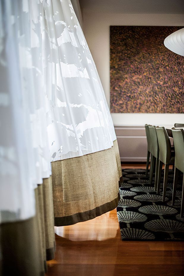 A styled dining room viewed through layered sheer and linen curtains. The room features a polished warm timber floor, a dark patterned carpet, a dining table with green upholstered chairs, a large textured brown wall artwork, and a white sculptural pendant light. The palette is warm and refined with strong patterned and organic textile elements.
