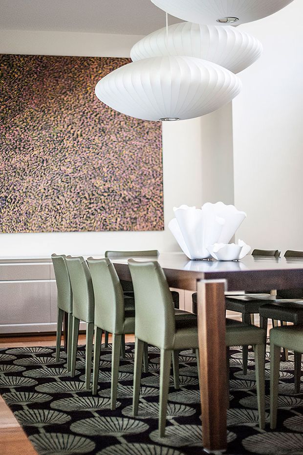 A refined dining room with a long dark wood dining table, upholstered sage-green dining chairs, sculptural white pendant lights, and a large abstract textured wall artwork. A bold patterned rug with fan-like motifs anchors the room, while a white sculptural vase forms the table centrepiece. The space has a retro-modern, polished character with strong graphic patterning.