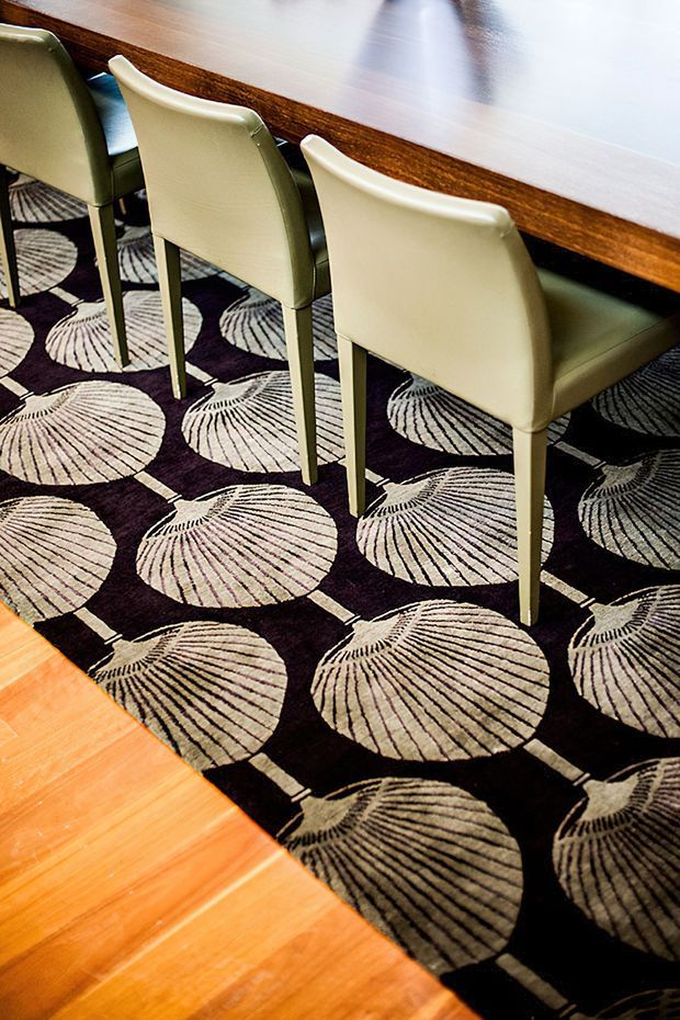 Close-up interior view of a dining area with a long dark timber dining table and several cream upholstered dining chairs. A bold black-and-beige patterned rug with large shell-like circular motifs sits over warm timber flooring.