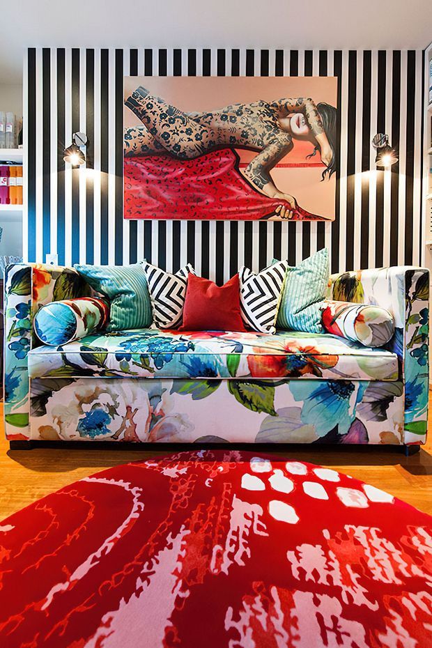 A vividly styled interior featuring a floral upholstered sofa with assorted patterned cushions, set against a dramatic black-and-white vertically striped feature wall. A large colourful artwork hangs above the sofa, while a bold red patterned rug covers the timber floor. The room has strong colour contrast, eclectic furnishings, and decorative wall sconces.