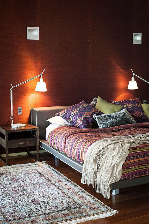 A richly styled bedroom with a low modern upholstered bed, dark reddish-brown textured wall finish, colourful geometric patterned bedding and cushions, a cream fringed throw, bedside table, and articulated reading lamps. A large distressed-pattern rug covers part of the dark timber floor, creating a warm, eclectic atmosphere.