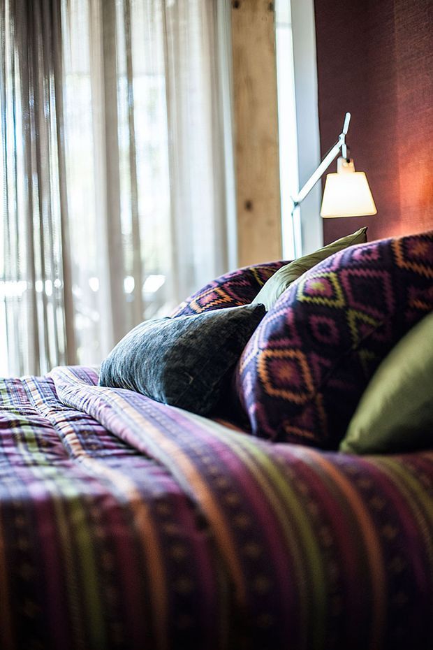 Close-up interior view of a richly patterned bed with layered multicoloured geometric bedding and cushions. A warm wall-mounted reading lamp sits against a deep burgundy feature wall, while sheer curtains diffuse daylight beside a natural timber structural post.