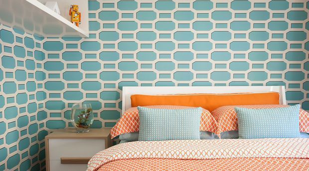 A colourful bedroom featuring a bed with an orange upholstered headboard, pale blue patterned pillows, and white bedding with orange geometric detailing. The room has a bold turquoise-and-white geometric feature wall, a white floating shelf with a small toy, and a bedside table with a small plant.