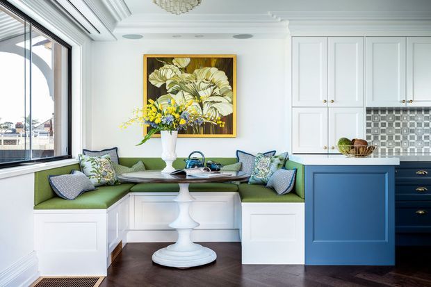 Bright, refined kitchen-dining nook featuring built-in green banquette seating, a white pedestal dining table, white panelled cabinetry, a deep blue island, patterned tile splashback, dark herringbone timber flooring, and a large window. The space has classic detailing with a fresh, colourful coastal-inspired interior.
