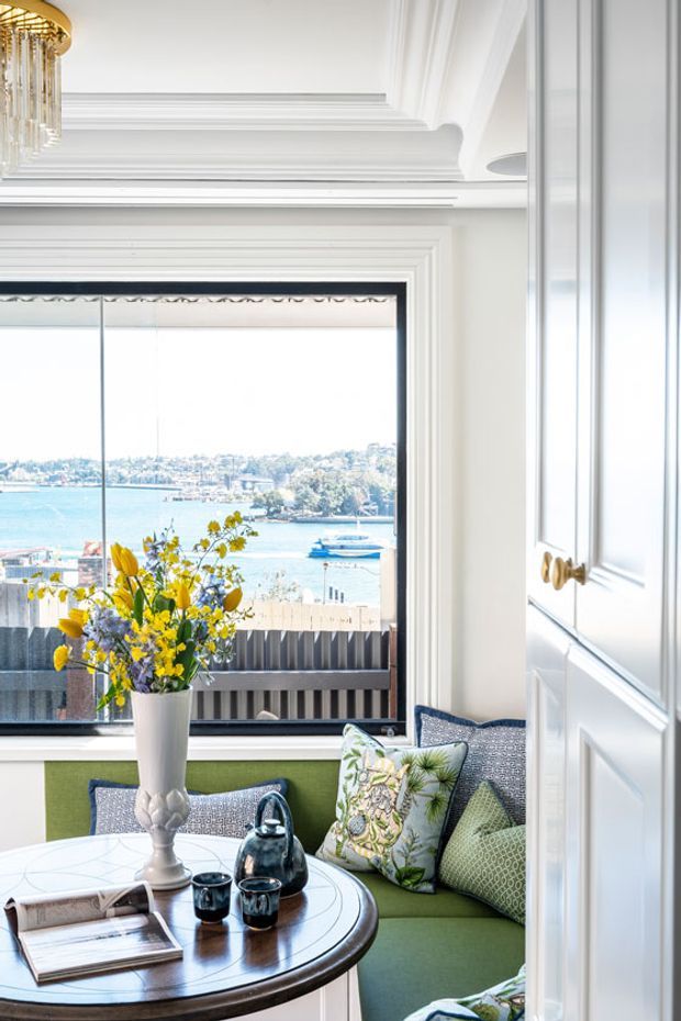 A bright, refined sitting area with a built-in green upholstered window seat, patterned cushions, a round dark timber table, and a floral arrangement. Large glazing overlooks a waterfront harbour, while ornate white wall panelling, layered cornices, and a decorative gold chandelier add a classic, elegant character.