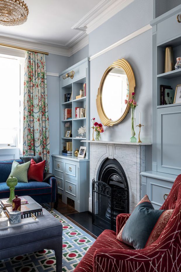 A refined, richly detailed living room with pale blue-grey panelled walls and built-in shelving, a marble fireplace with black cast-iron insert, and an ornate oval gold-framed mirror. The room contains a deep blue sofa, a red patterned armchair, an upholstered ottoman, colourful geometric rug, floral curtains, decorative objects, and an ornate crystal chandelier. The overall composition is colourful, layered, and traditionally styled with strong period-inspired detailing.