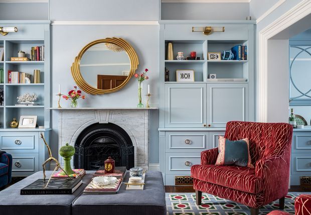 A refined living room with pale blue built-in shelving and cabinetry surrounding a traditional fireplace. A large ornate gold round mirror hangs above the mantel, with a deep red patterned armchair, dark upholstered ottoman, books, decorative objects, and a colourful geometric rug adding rich accents. The room combines classic architectural detailing with layered, elegant furnishings.