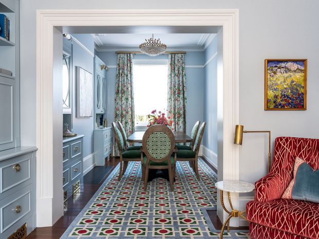 A refined, traditionally styled dining room viewed through a wide trimmed doorway from an adjoining sitting area. The room features pale blue panelled walls, dark timber flooring, a patterned area rug, a central round dining table with upholstered chairs, floral curtains, decorative cabinetry, and an ornate chandelier. A red patterned armchair and small side table occupy the foreground.