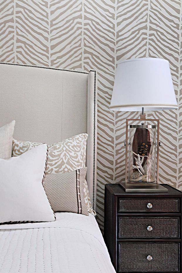 Close-up of a refined bedroom vignette featuring a tall upholstered headboard with nailhead trim, layered white and patterned bedding, a dark textured bedside chest, and a table lamp with a decorative glass base. The walls are covered in a bold beige-and-off-white organic leaf or zebra-inspired patterned wallpaper.