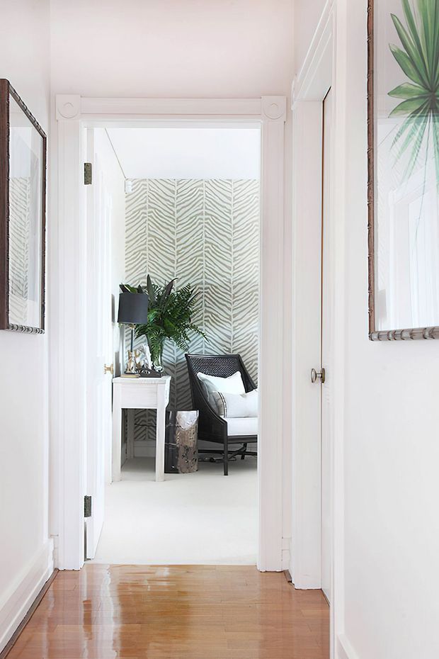 Bright, narrow residential hallway with glossy timber flooring, white walls, decorative white door and window trim, and framed botanical artwork. An open doorway leads into a small sitting area featuring a dark woven armchair, white cushions, a side table, lamp, potted greenery, and a bold zebra-patterned wallpaper feature wall.