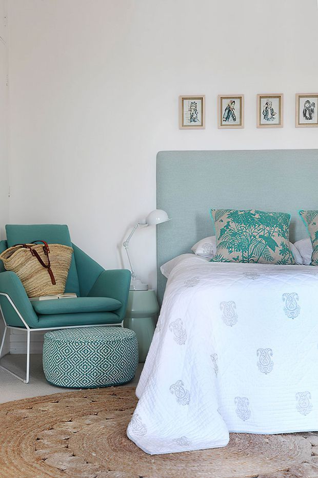 A bright, softly styled bedroom with a pale aqua upholstered bedhead, white bedding with subtle printed motifs, and a matching teal armchair beside a patterned round ottoman. A small bedside table and white articulated lamp sit between the chair and bed, while a row of framed illustrations decorates the white wall. A natural woven rug adds texture underfoot.