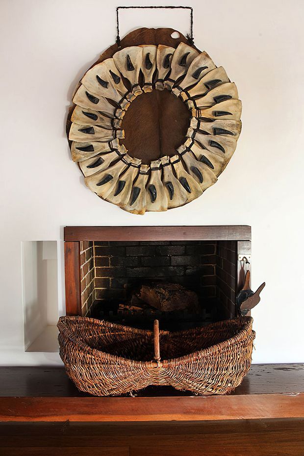A close-up interior view of a traditional fireplace with a dark brick firebox and timber mantel trim. A large woven wicker log basket sits in front of the hearth, with stacked firewood inside. Above the fireplace is a distinctive circular decorative wall piece made from curved natural materials. The room has white plaster walls and a dark timber floor and hearth surround.