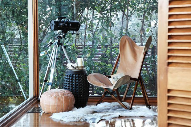 A cozy, light-filled glazed corner furnished as a reading and relaxation nook. Floor-to-ceiling windows overlook dense greenery, with a vintage telescope on a tripod, a leather sling chair, a small woven side table, a leather pouf, and a sheepskin rug on polished timber flooring. Timber-framed glazing and a timber louvred screen add warmth and texture.