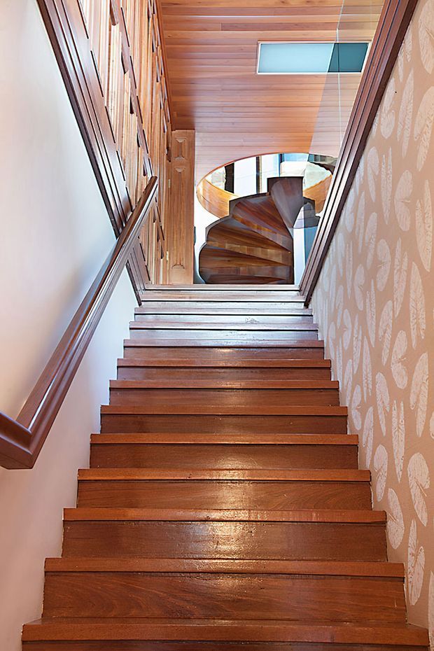 Interior view looking up a narrow timber staircase toward a landing and distinctive curved spiral stair beyond. The stair treads, wall panelling, ceiling boards, and trim are finished in warm polished wood, with patterned wallpaper on the right wall, a skylight above, and strong natural light from glazing at the upper level.