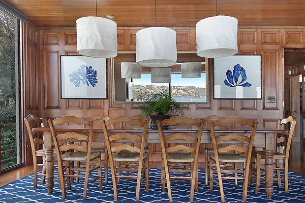 A richly detailed dining room with a long timber dining table and matching carved wooden chairs arranged beneath oversized white pendant lights. The walls feature dark timber paneling with integrated framed artwork and a large central mirror or glazed opening, while a blue patterned rug adds contrast. The room has a warm, refined, traditional character with strong timber detailing.