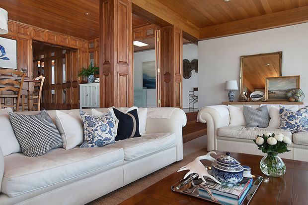 Traditional living room with white upholstered sofas, patterned cushions, a wooden coffee table, decorative accessories, and extensive dark timber wall panelling, columns, door frames, and ceiling. A dining area and hallway are visible in the background, with a large wall mirror and artwork adding depth.