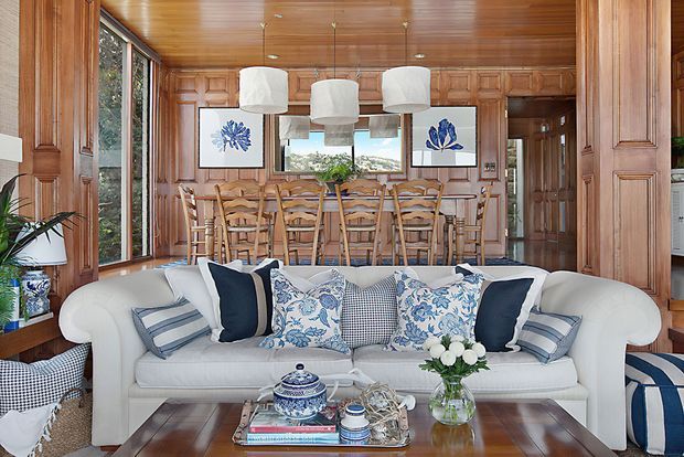 A richly detailed open-plan living and dining area with a white upholstered sofa in the foreground and a timber dining table with woven chairs behind it. The room features extensive warm timber wall panelling and ceiling boards, built-in cabinetry, framed blue-and-white botanical artwork, patterned blue-and-white cushions, and multiple white pendant lights. Large windows bring in natural light and views of greenery, creating a refined coastal-traditional atmosphere.