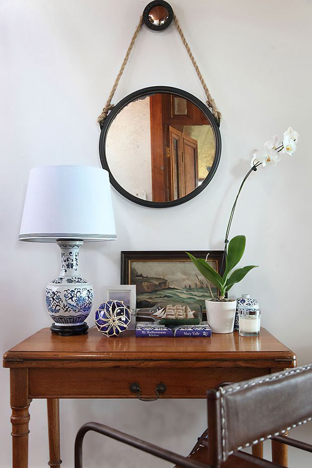 A styled console or writing desk vignette against a white wall, featuring a dark timber desk with turned legs and drawer, a blue-and-white ceramic table lamp, decorative objects, framed artwork, and layered round mirrors including a rope-hung mirror. A leather chair is partially visible in the foreground. The composition has a refined, collected traditional character.