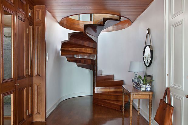 Interior view of a refined entry hall featuring a sculptural dark timber spiral staircase, matching timber-paneled ceiling, polished timber flooring, white paneled walls and doors, and traditional built-in cabinetry. A console table, mirror, lamp, and decorative accessories complete the space.