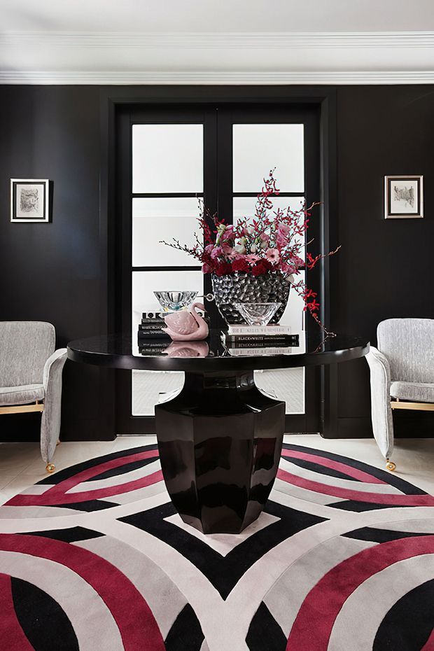 A dramatic, symmetrical interior vignette featuring a glossy black pedestal table in front of a black-framed glazed door or window. Two pale upholstered armchairs flank the table, which is styled with stacked books, glassware, a pink decorative vessel, and a large floral arrangement. A bold geometric rug in black, white, grey, and deep red covers the floor. Black walls are contrasted with white decorative ceiling cornices and trim, creating a polished, theatrical setting.