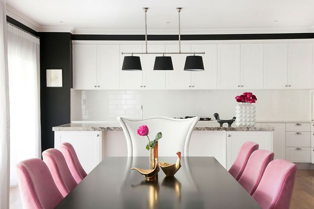 A bright, refined open-plan dining area adjoining a white kitchen. The dining table has a dark glossy surface and is surrounded by sculptural blush-pink upholstered chairs, with brass decorative objects and a flower arrangement as accents. The kitchen features extensive white shaker-style cabinetry, a pale stone benchtop, white subway-tile splashback, black trim and pendant lights, and a dark contrasting wall.