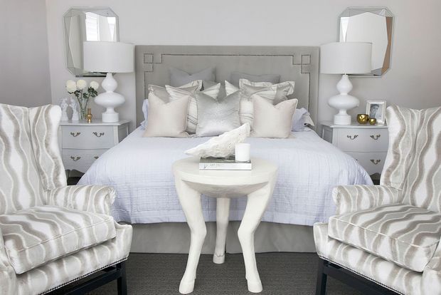 A symmetrical, softly styled bedroom with a pale upholstered bed and layered white and grey bedding, flanked by matching bedside tables, white lamps, and geometric wall mirrors. Two patterned accent armchairs frame a small sculptural white table in the foreground. The overall design is refined, light, and neutral with subtle metallic and grey details.