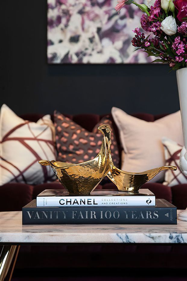 A styled interior vignette featuring two polished gold bird sculptures displayed on stacked fashion and design books atop a marble coffee table. A deep charcoal wall, abstract artwork, burgundy seating, patterned cushions, and a tall vase of pink and white flowers create a dramatic, luxurious composition.