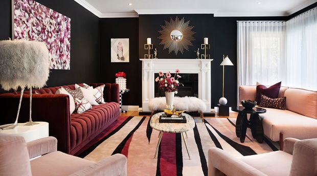 Luxurious, highly styled living room with dramatic black walls, white trim and fireplace surround, velvet seating in burgundy and blush tones, a bold geometric striped rug, statement sunburst mirror, sculptural lighting, and layered floral and metallic accents. Large sheer-curtained windows bring in soft daylight.