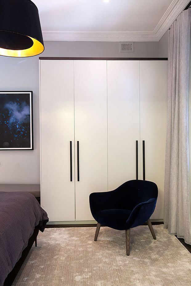 A refined bedroom interior featuring a full-height white built-in wardrobe with long black handles, a dark upholstered accent chair, a partially visible bed, a neutral textured rug, floor-to-ceiling curtains, and decorative ceiling cornices. A black pendant light with a warm inner glow hangs at the upper left, while framed artwork adds a dark blue accent.