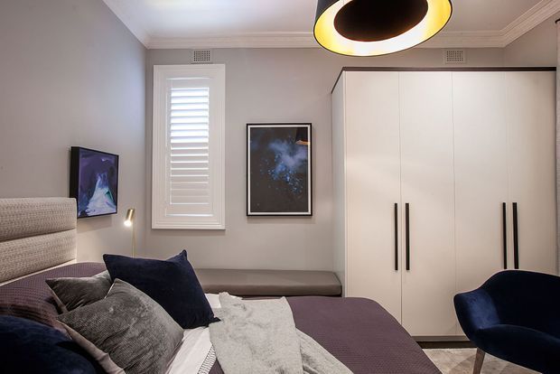 A compact, refined bedroom with a large upholstered bed in the foreground, deep purple and grey bedding, a navy accent chair, and full-height white built-in wardrobes with black vertical handles. The room has pale grey walls, white plantation shutters, decorative wall art, a sculptural circular pendant light, and classic ceiling cornices.
