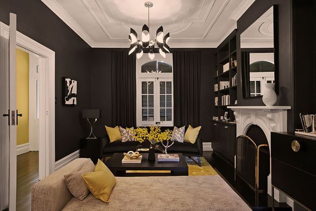 A sophisticated dark-toned living room with black walls, ornate white ceiling detailing and mouldings, a contemporary chandelier, built-in black shelving, and a white decorative fireplace. A black sofa with yellow and patterned cushions faces a low coffee table, while a large upholstered foreground piece adds softness. Shuttered windows and a bright adjoining hallway are visible.