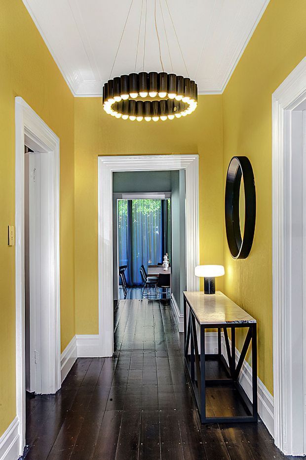 A narrow, brightly styled interior hallway with mustard-yellow walls, dark timber flooring, white decorative trim and a white paneled ceiling. A large circular pendant light hangs centrally, while a slim dark console table with a lamp and round black-framed mirror lines the right wall. White-trimmed doorways lead to adjacent rooms, including a visible dining or workspace area at the far end.