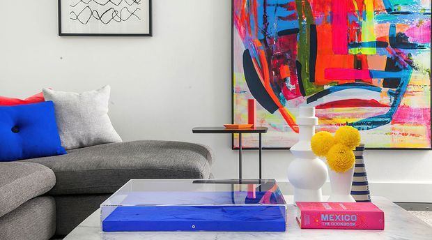A contemporary living room with a large grey sectional sofa, vivid blue and red accent cushions, a transparent glass coffee table with a bold blue base, and colourful abstract artwork. Sculptural white vases, yellow flowers, and a small side table add decorative accents to the bright, eclectic setting.