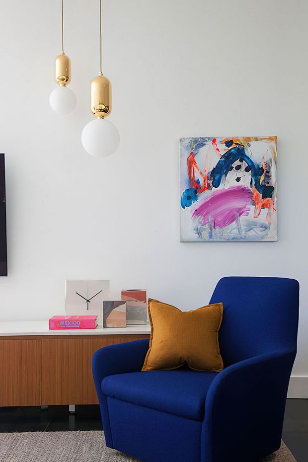 A stylish modern living room vignette with a deep blue upholstered armchair, mustard-gold cushion, low timber sideboard, colorful abstract artwork, and two sculptural pendant lights with cream and brass-toned shades. The room has pale grey walls, dark flooring, and a textured neutral rug.