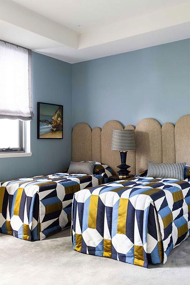 A compact twin bedroom with two single beds dressed in bold geometric quilts in navy, blue, mustard yellow, white, and grey. Each bed has a tall rounded upholstered headboard in taupe-beige, with patterned cushions and a central table lamp. The room has pale blue walls, a white ceiling, a carpeted floor, a curtained window, and a framed artwork. The overall look is colourful, playful, and tailored.
